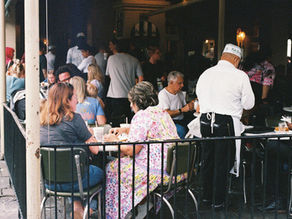 People sit and chat at outdoor café tables. A waiter in white uniform serves drinks. The café is bustling with activity and conversation.