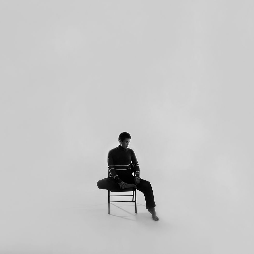 Black-and-white portrait of a man in a dark turtleneck sitting on a chair in a stark white studio, looking down calmly.