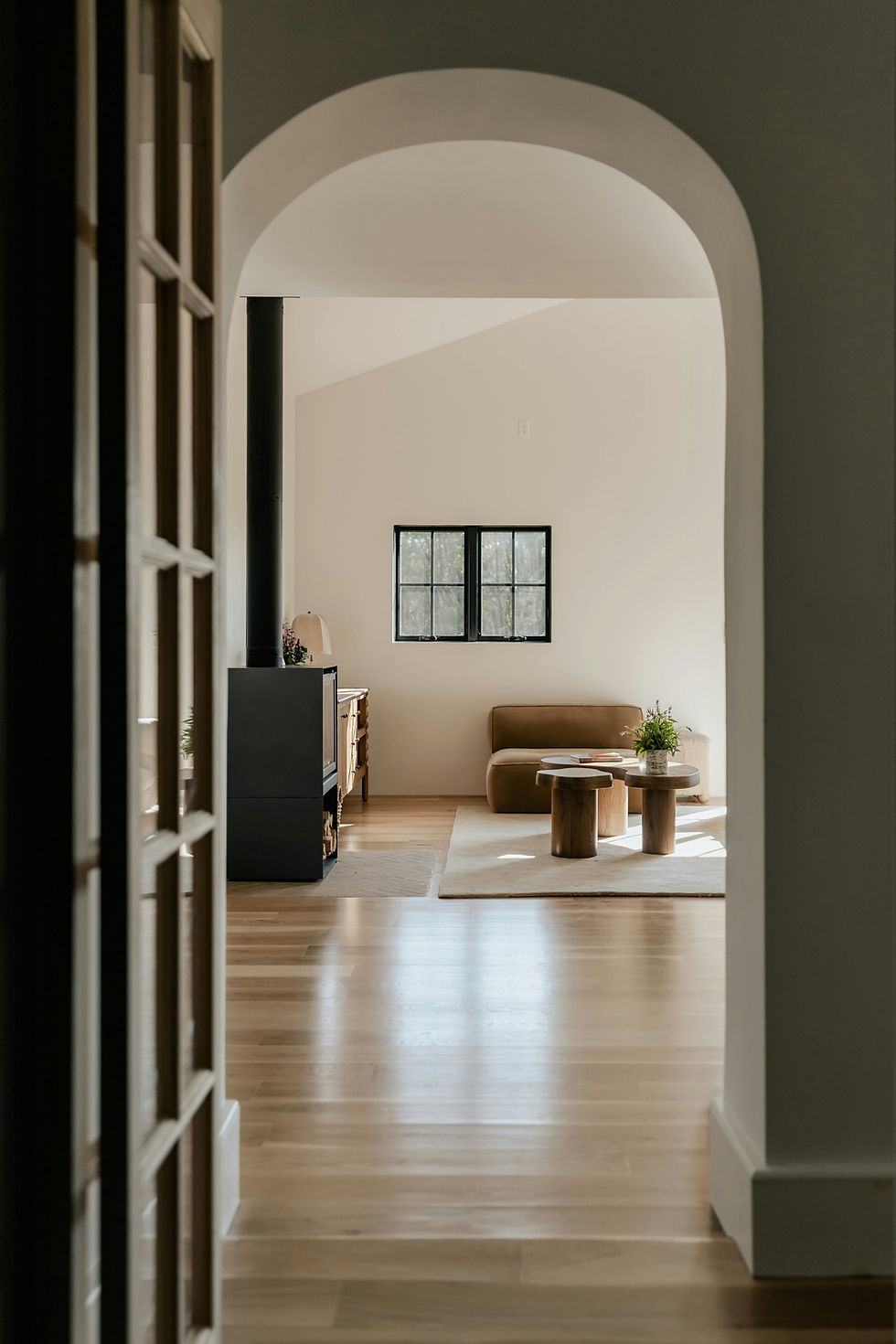 Bright living room with arched doorway, beige walls, wooden floor, brown sofa, wooden table, and window. Cozy, minimalist atmosphere.
