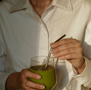 Person in a white shirt stirs a green smoothie in a glass with a metal straw, lit by warm sunlight.