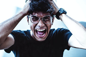 Man in glasses screams with hands on his head, wearing a black shirt and watch against a bright white-blue background.