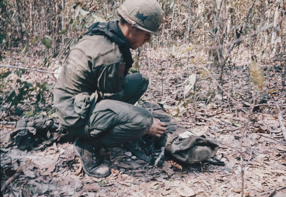 A soldier in uniform and helmet is crouched in a forest, examining gear on the ground. The scene is colored in earthy tones.