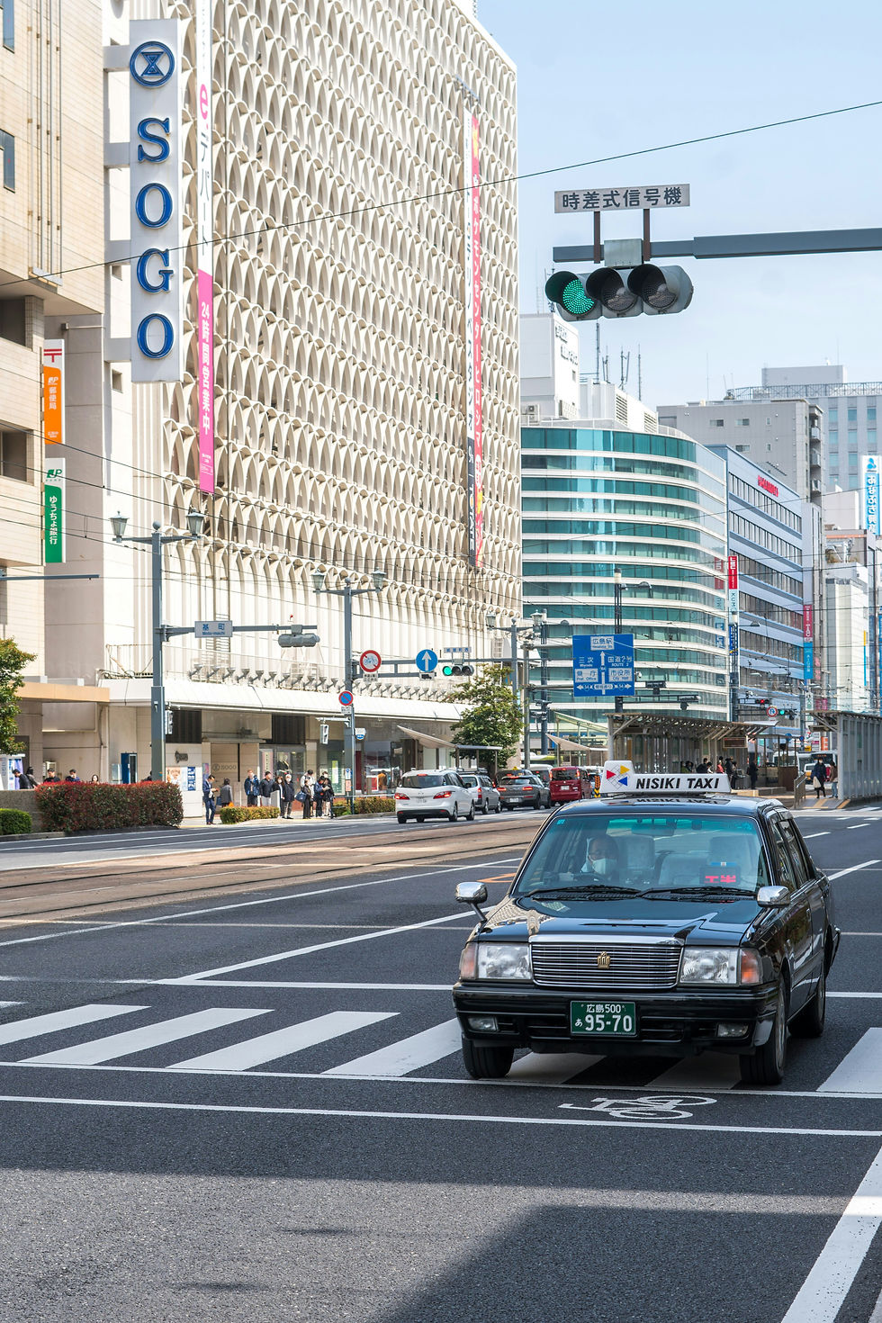 Taxi at crosswalk in front of SOGO building on a busy city street. People walk nearby, with modern buildings in the background.