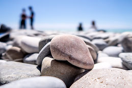 Close-up of smooth pebbles on a shore with a blurred background of people standing and a blue sky.