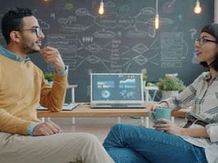 Zwei Menschen unterhalten sich in einem modernen Büro. Ein Laptop mit Diagrammen steht vor ihnen. Hintergrund: Tafel mit Skizzen.