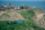 Man in jeans lying on grassy cliff under a blue sky by the sea. Houses and people on a path in the background. Peaceful scene.