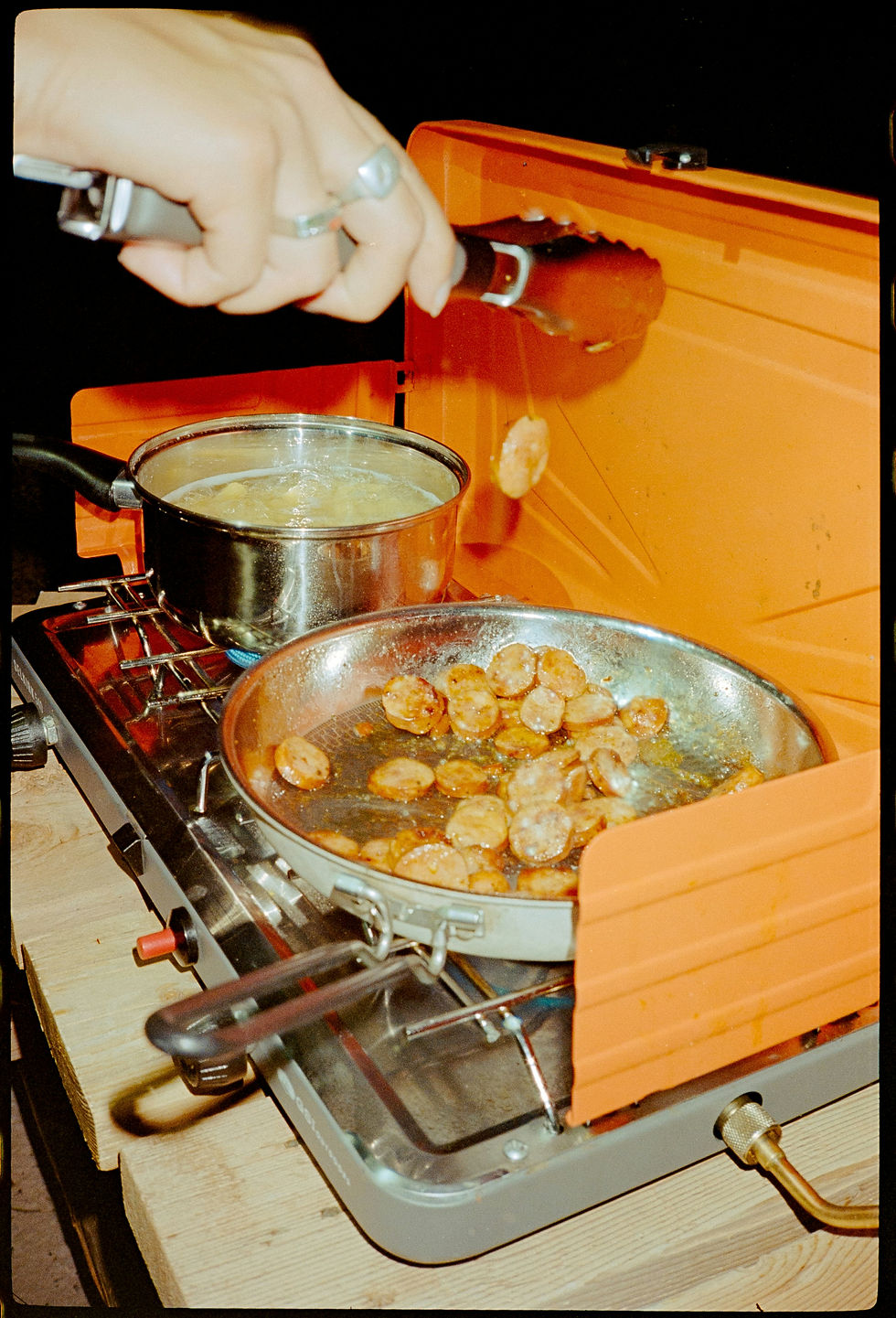 Hand with tongs cooks sausage on an orange camping stove beside a boiling pot and pan of browned slices
