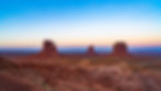 Stock image of Three majestic buttes in Monument Valley at sunrise, with vibrant red desert terrain and a clear blue to pink gradient sky.