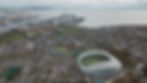 Aerial view of Dublin's Aviva Stadium, surrounded by cityscape and coastline. Overcast sky, stadium text visible: AVIVA. Calm atmosphere.