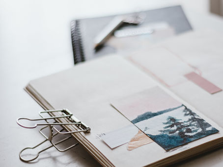 Open notebook with nature-themed collage and paper clip on a light wooden table. Another notebook and pen slightly blurred in the background.