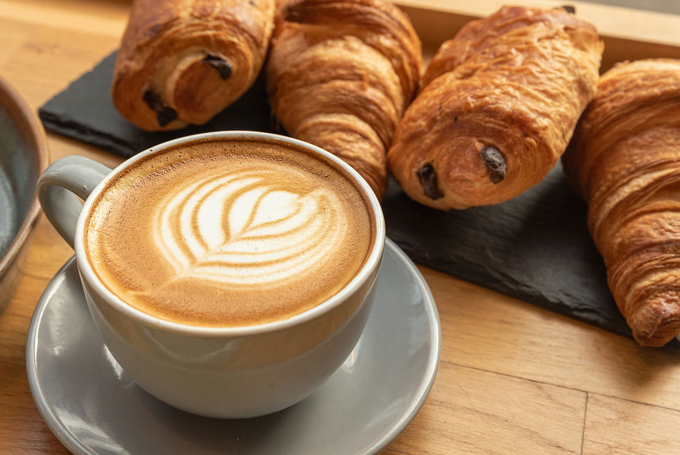 Latte with leaf foam art in a gray cup beside four golden croissants on a black slate board on a wooden table