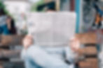 medium close up image of a person in a striped shirt sitting on a bench, holding up a newspaper in front of their face