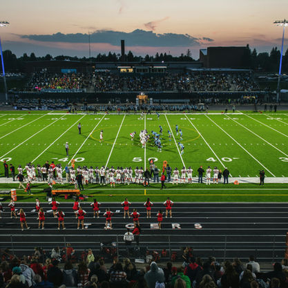 Football game at sunset with teams on bright green field, cheerleaders and crowd on sidelines, stadium lights illuminating the scene.