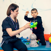Therapist and young boy play with colorful toys on a padded mat in a playroom; boy wears an ALWAYS sweatshirt.