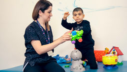 Therapist and young boy play with colorful toys on a padded mat in a playroom; boy wears an ALWAYS sweatshirt.