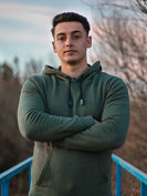 Young man in green hoodie with arms crossed, trees in background.