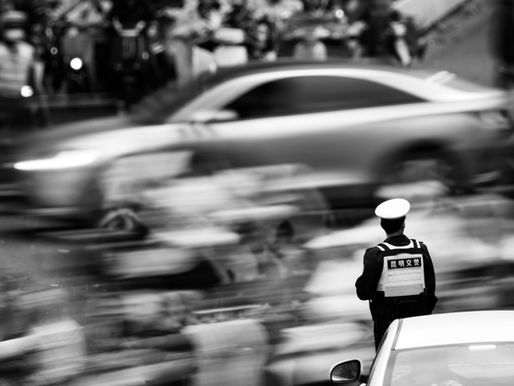 Police officer in uniform stands in a busy, blurred street scene with passing cars and pedestrians. Black and white image captures motion.
