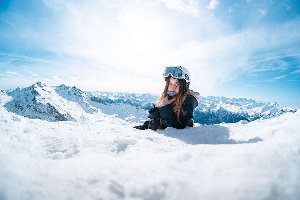 A girl at the top of the mountain wearing snowboarding gear, the sun behind her.