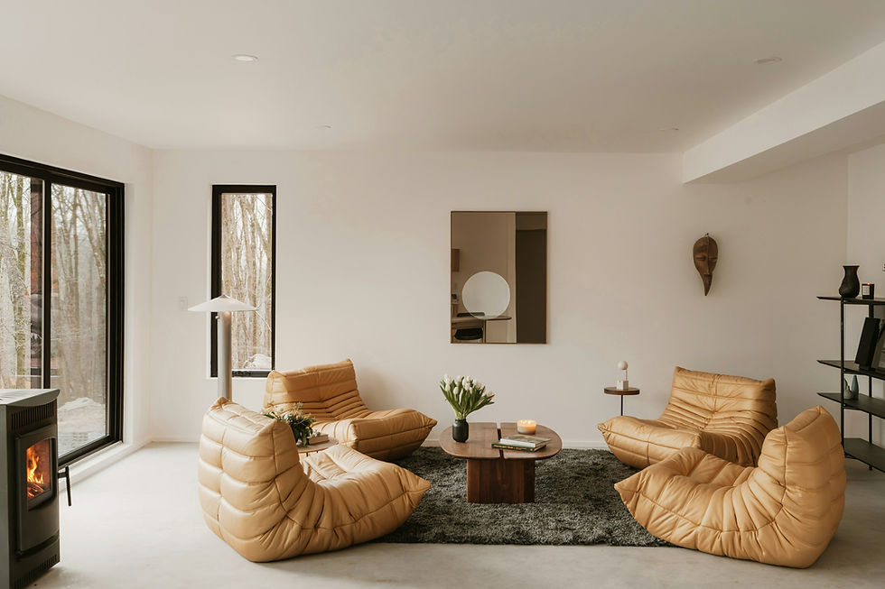 Cozy living room with beige chairs, a wooden table, and tulips. Large windows show a forest view. Modern decor with a lit fireplace.