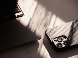 Two smartphones on a sunlit table, one matte black and one carbon-fiber, with soft window shadows.
