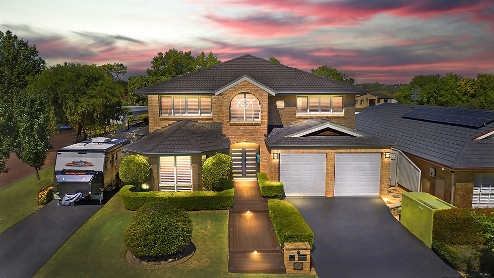 Two-story brick house at sunset with lit windows, double garage, caravan in driveway, and a colorful pink-purple sky.