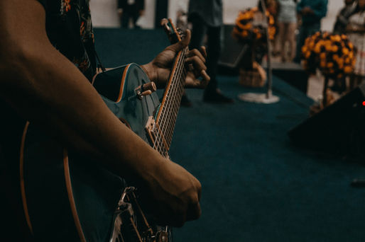 Close-up of a person playing a teal electric guitar. In the blurred background, people stand near orange flower arrangements. Indoor setting, moody tone.