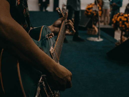 Close-up of a person playing a teal electric guitar. In the blurred background, people stand near orange flower arrangements. Indoor setting, moody tone.