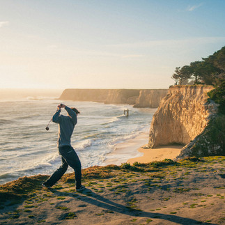 man swinging a golf club on the hillside next to the ocean 
