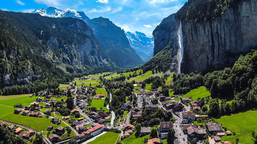 Aerial view of a valley with a village, green fields, and a waterfall cascading down a cliff. Snow-capped mountains in the background, clear sky.