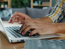 Hands typing on a laptop at a wooden desk in a cozy living room, with blurred shelves and cushions in back.