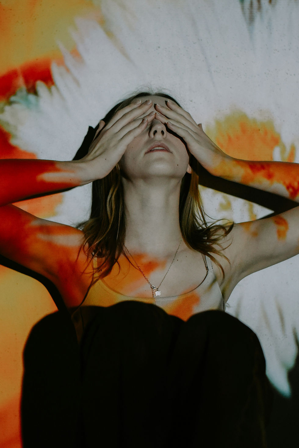 Woman covering her eyes with hands, sitting against a wall with vibrant red, orange, and white abstract projection. Reflective, introspective mood.