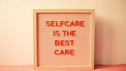 Wood-framed sign with red text "SELFCARE IS THE BEST CARE" on pink background. Set against a cream wall. Calm, minimalist vibe.