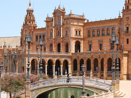 Ornate building with arched facades, a tiled bridge, and lampposts. Two people stand on the bridge, surrounded by blooming trees. Bright day.
