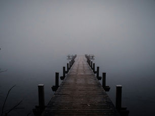 Wooden pier extends into foggy, serene lake. Bare branches frame the edges. Moody, muted blues and grays dominate the misty scene.