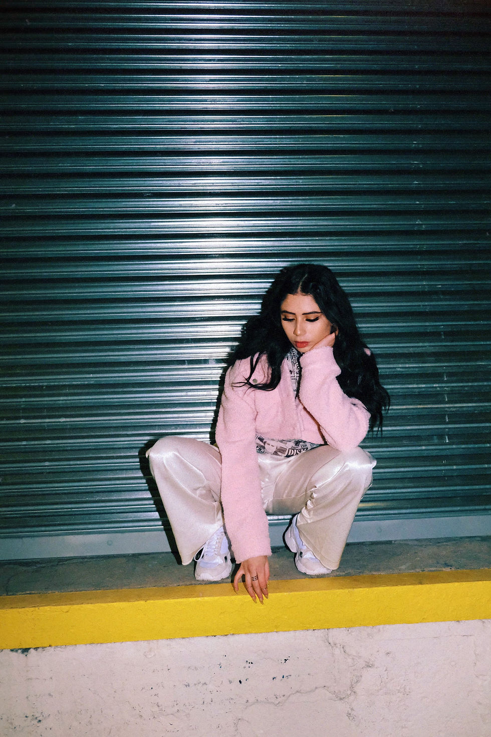 Young woman in a pink jacket crouches on a yellow ledge against a green metal shutter. She looks thoughtful, with a hand on her face.