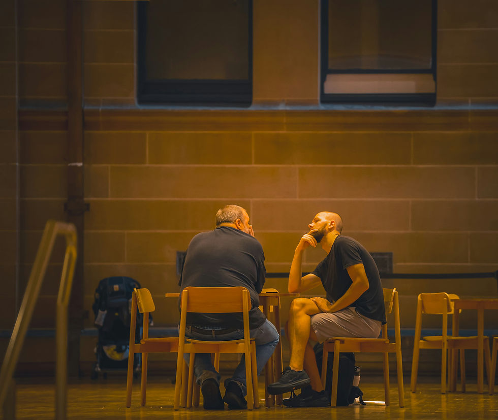 Two men sit at a table in deep conversation in a dimly lit room with brown walls. One man rests his chin on his hand, appearing thoughtful.