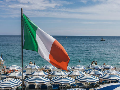 Italian flag waves above a beach with blue-striped umbrellas and sunbathers. Calm sea and a distant boat under a clear sky.