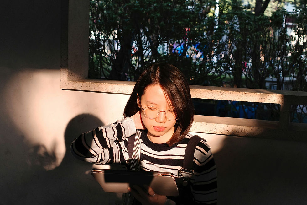 A young female student wearing glasses and a striped sweater, sitting in a sunlit area while thoughtfully looking down at a tablet.