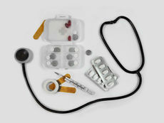Ein Stethoskop liegt auf einem weißen Hintergrund neben verschiedenen Medikamentenblistern, einer Tablettenbox, einem Fieberthermometer, einem Verband und medizinischem Klebeband. Die Anordnung symbolisiert medizinische Versorgung, Diagnostik und Behandlung.