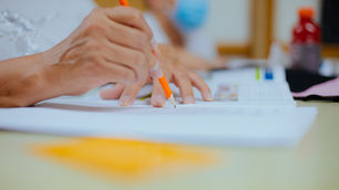 Hands writing with an orange pencil on paper, in a classroom. A person with a blue mask is in the background, creating a focused atmosphere.