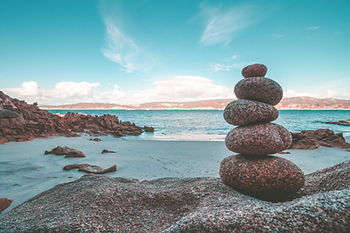 Stack of smooth stones on a sandy beach, with an ocean and rocky shoreline in the background. Sky is bright blue with wispy clouds.