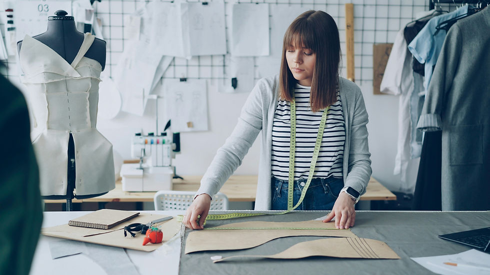 A woman measures fabric with a tape in a sewing room. Mannequin and sewing tools are visible. Papers and patterns cover the wall.