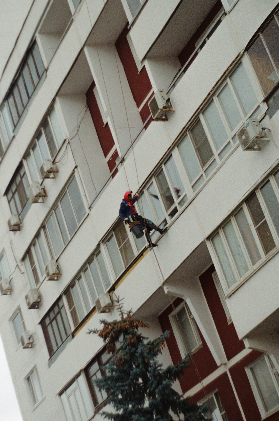 A worker in safety gear cleans windows on a tall building. The facade is red and white, with air conditioners visible. A tree is below.