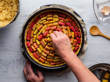 Hands arranging rhubarb pieces in a tart on a light wooden table. Nearby, a bowl with dough, a wooden spoon, and cracked eggshells.