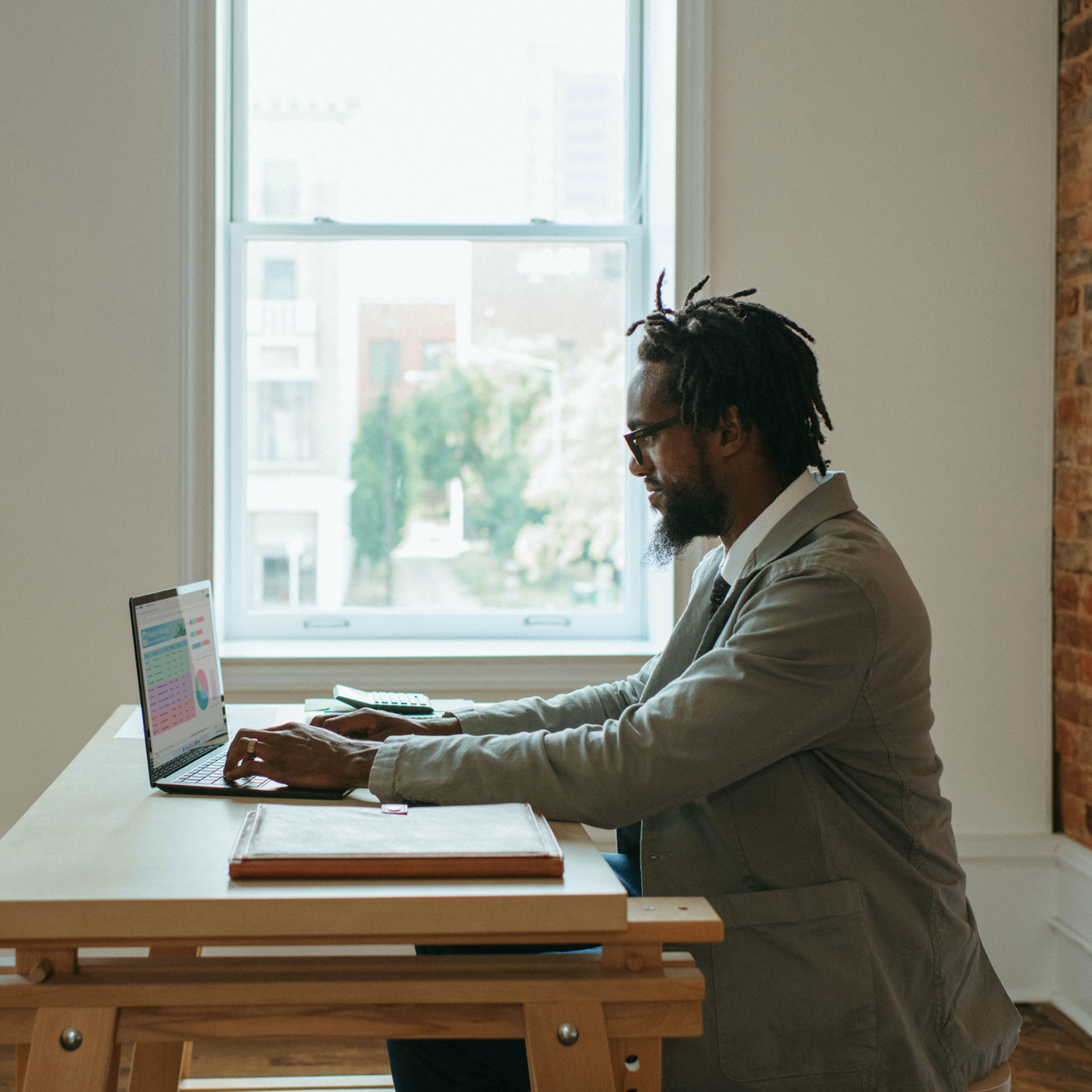 Man in a suit works on a laptop at a wooden table in a bright room with an exposed brick wall and large window showing a city view.