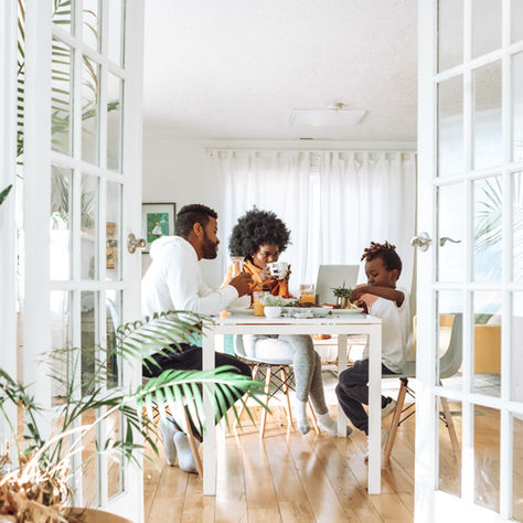 Family having breakfast at a white table in a bright room. Plants and glass doors surround them, creating a cozy, relaxed atmosphere.