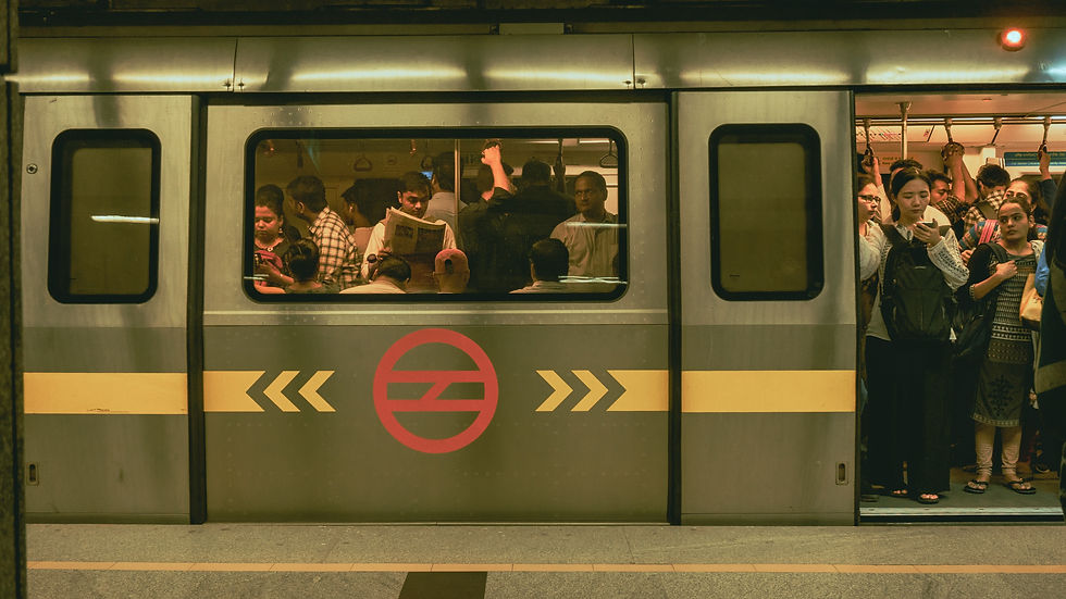 Crowded metro train with passengers standing, reading, and using phones. Gray exterior with a red logo. Dim station lighting adds a warm tone.