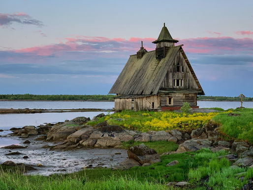 A wooden house on rocky shore, surrounded by vibrant green grass and yellow flowers, with a serene lake and pink-tinged evening sky.