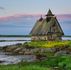 A wooden house on rocky shore, surrounded by vibrant green grass and yellow flowers, with a serene lake and pink-tinged evening sky.