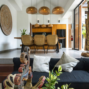 Modern living room with a black sofa, patterned wood sculpture, and plants. Woven chairs around a wooden table under pendant lights. Cozy and stylish.